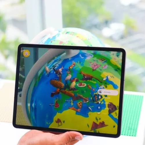 Shifu Orboot AR globe with tablet showing AR animals over a colorful world map, held in hand on table in front of window.